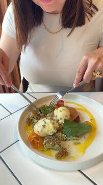 young woman having breakfast or brunch in a cozy cafe, cutting zucchini pancakes topped with stracciatella cheese and fresh vegetables