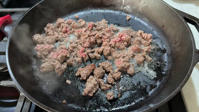 Close up 4K video of ground beef sizzling and browning in a cast iron skillet on a home stovetop. 