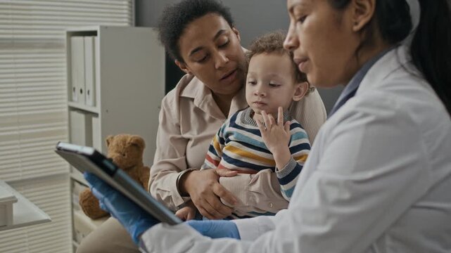 Hispanic female doctor showing medical scans on tablet to biracial mother holding young child. Footage ideal for pediatric clinic advertising and digital healthcare services
