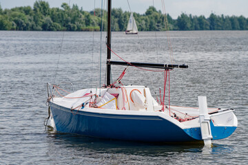 A small blue yacht with lowered sails rests peacefully on tranquil water. Green trees line the distant shore. Ideal for nautical themes and summer vacation backgrounds. Photo © slexp880