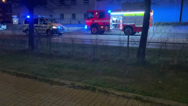 Night police car and fire engine parked with flashing lights, officer silhouette near patrol vehicle and crew checking scene, blue and red lights