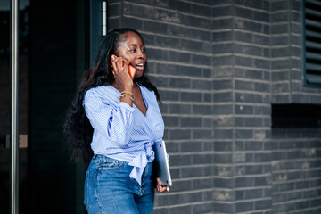 Fototapeta premium Black woman talking on phone carrying laptop