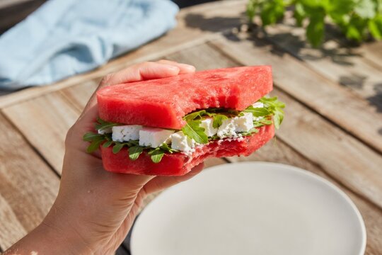 Hand holding a creative watermelon sandwich with feta cheese and arugula, showing a big bite, outdoors on a sunny day. A healthy, refreshing summer snack concept on a rustic wooden table.