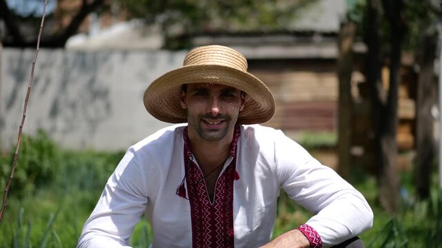 Portrait of ukrainian man in straw hat against background of summer garden. Young smiling guy wearing an embroidered shirt looking into camera outdoor. Concept of national identity and patriotism