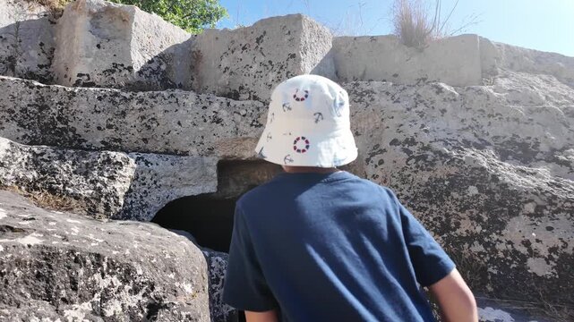 Salina, Malta - a young boy stands before a dark, rectangular entrance carved into weathered limestone rock, marking one of the ancient Paleo-Christian catacombs surrounded by layered stone blocks.