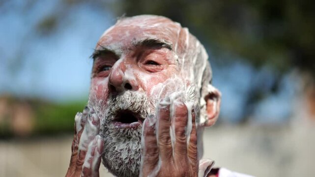 Elderly man wearing in traditional embroidered shirt washing his gray beard fully covered in soap at summer garden. Scene captures care with sense of peace and natural simplicity. Daily ritual concept