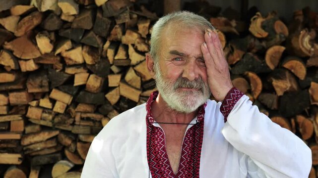 Senior ukrainian man in vyshyvanka standing after hard work near woodshed. Elder tired male in straw hat resting after hard job near firewood. Concept of national identity and independence. Close up
