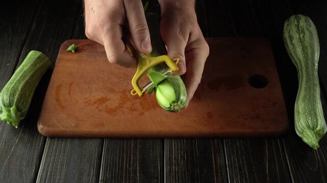 Hands peeling zucchini with a yellow vegetable peeler on a wooden cutting board, surrounded by fresh zucchinis in a rustic kitchen setting