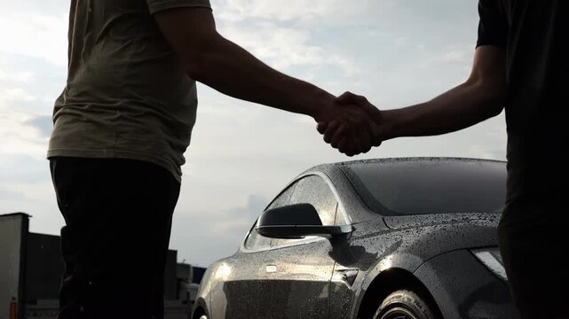 Male arm giving key of car to buyer of electric automobile at rainy day. Handshake between salesman and new owner of modern auto after successful deal at sunset. Concept of buy or rent vehicle