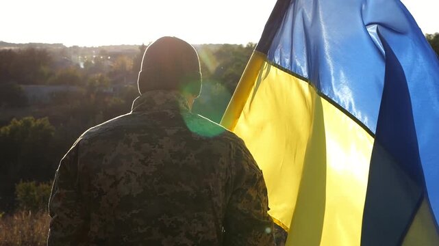 Male soldier in military uniform with Ukraine flag as symbol of victory against russian aggression. Ukrainian army man stands with national banner at countryside. Concept of invasion resistance
