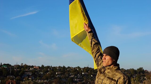 Ukrainian army man stands with raised national banner to peak of hill. Male soldier in military uniform with Ukraine flag as symbol of victory against russian aggression. Invasion resistance concept