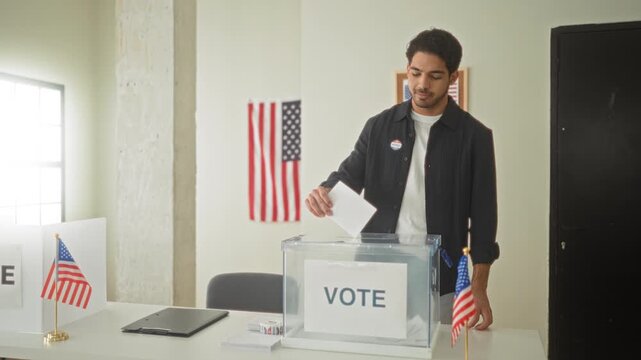 Young hispanic man inserting ballot with his hand into a clear ballot box at a polling building with american flags visible; civic duty pride.