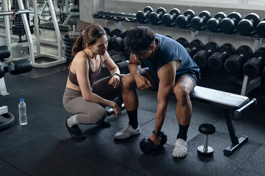 Woman Checking on Tired Man Resting on Bench Beside Dumbbell Rack.