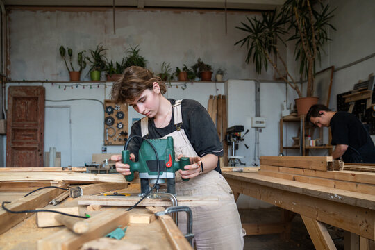 Teamwork and craftsmanship in restoration workshop, woman crafter processing board with wood router, focused female worker trimming and carving surface of wooden plank with plunge router at workbench.