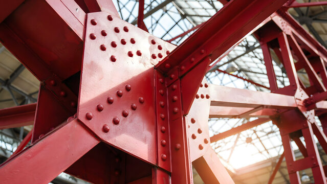 Red painted steel girder structure with visible rivets and bolts inside a building with a glass roof and natural light shining through the beams
