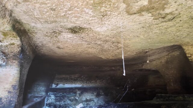 Salina, Malta - a detail shot pans across the weathered stone ceiling of an ancient Paleo-Christian catacomb, where hanging spider webs and moss reveal the age and stillness of the underground chamber