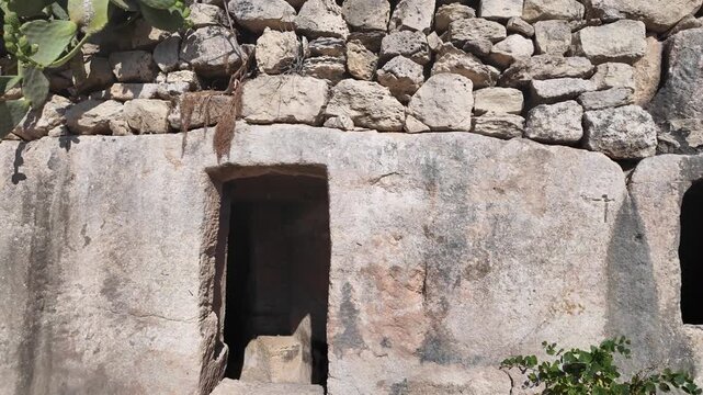 Salina, Malta - a panning shot reveals a row of ancient Paleo-Christian catacomb entrances carved into limestone rock, framed by dry stone walls and trailing ivy under bright sunlight.