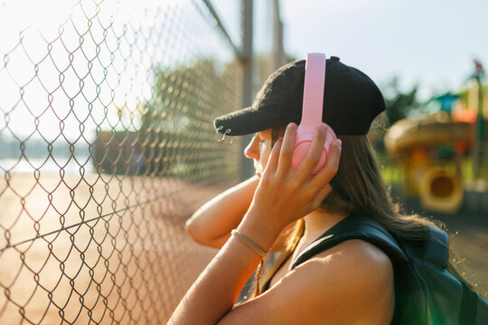 Close up profile of a teenage girl adjusting pink headphones near a sports fence.