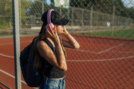 Profile of female student holding pink headphones by a wire fence at sunset.