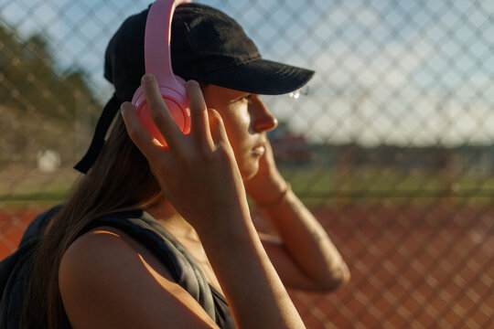Close up profile of a teenage girl holding pink headphones by a sports fence.