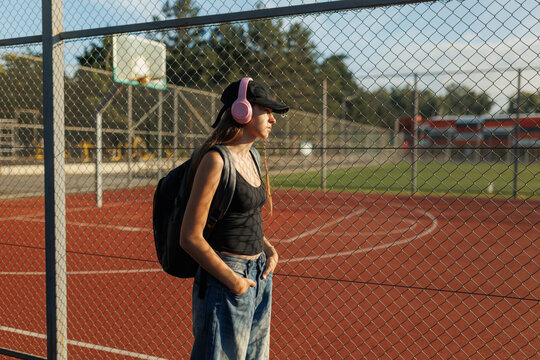 Teenage girl with pink headphones looking through a sports fence at sunset.