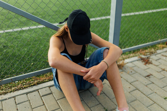 Depressed teenage girl sitting on pavement hiding face near a sports fence.