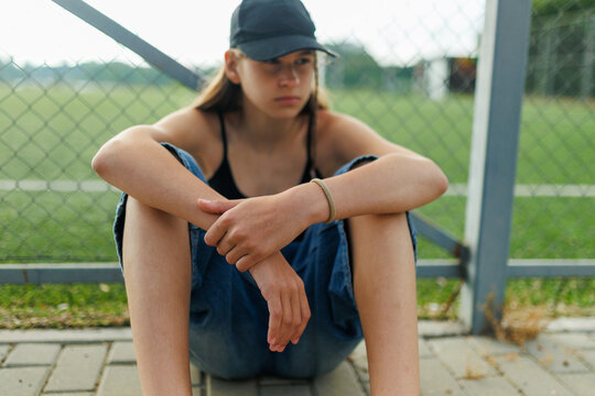 Sad teenage girl sitting on pavement by a sports fence during late afternoon.