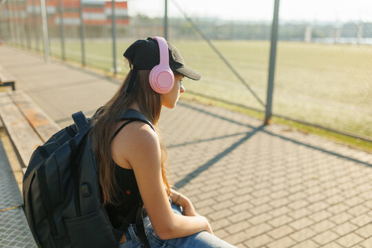 Teenage girl with pink headphones and backpack sitting near a sports fence.