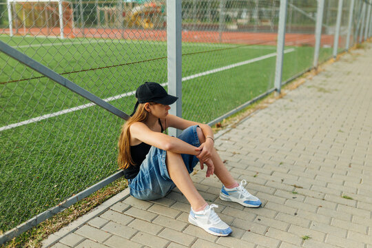 Lonely teenage girl sitting on pavement leaning against an outdoor sports fence.