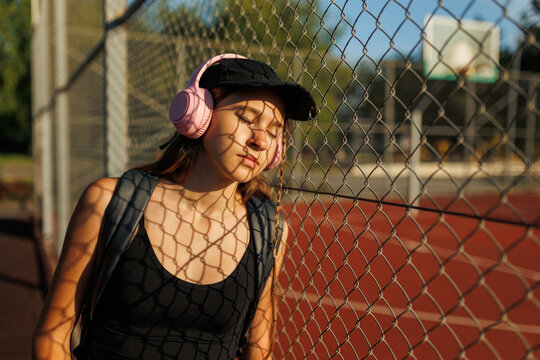 Teenage girl with pink headphones resting on a wire sports fence at sunset.