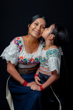 Indigenous Kichwa mother and daughter from Otavalo, Ecuador, wearing traditional hand-embroidered blouses and anaco sharing affectionate moments of cultural heritage in a studio portrait