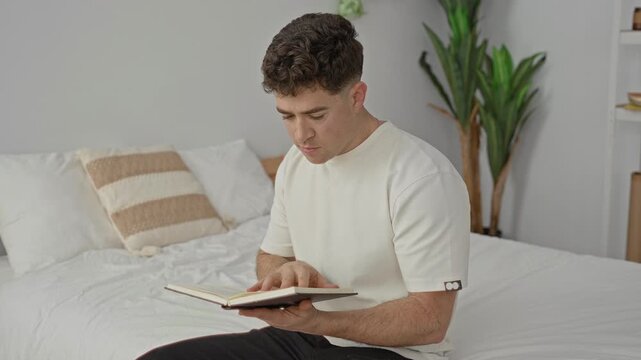 Man reading book on bed, hand turning page, seated on white bedding with striped pillow and plant, set in studio; calm contemplation.