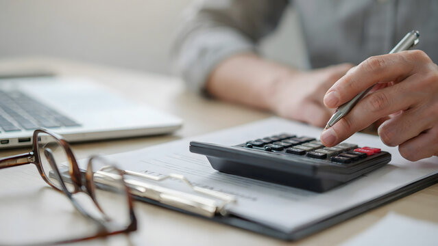 A person in a grey shirt using a black calculator and writing on a document at a wooden desk with reading glasses resting on the table