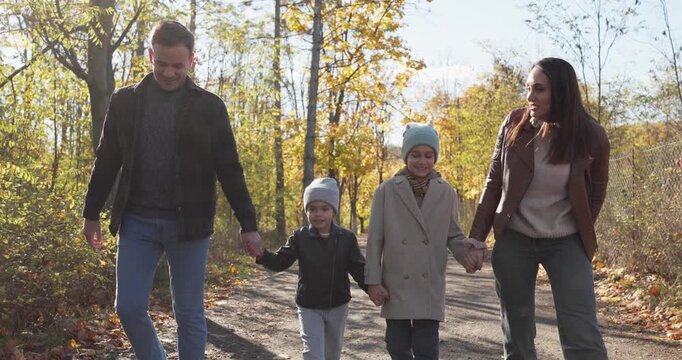 Happy family walking on path through autumn park with carefree smiles and talks. Cheerful father, mother and kids wearing jackets and hats enjoy weekend walk, parents and children relax together