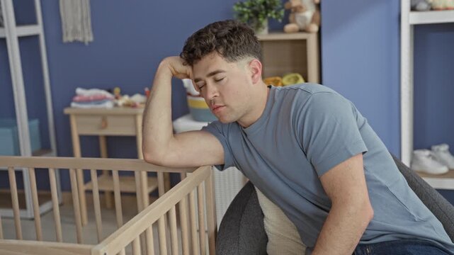 Man seated with forearm draped over wooden crib rail, eyes closed, leaning back in a blue nursery building; quiet exhaustion parenting struggle.