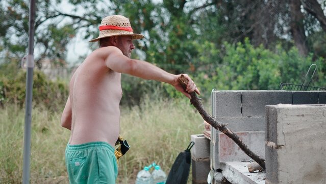 Man in a straw hat feeding wood to an outdoor paella fire