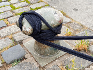 Weathered metal mooring bollard secured with thick black rope on cobblestone pavement, surrounded by patches of grass and concrete © Vlad Yakubovskiy