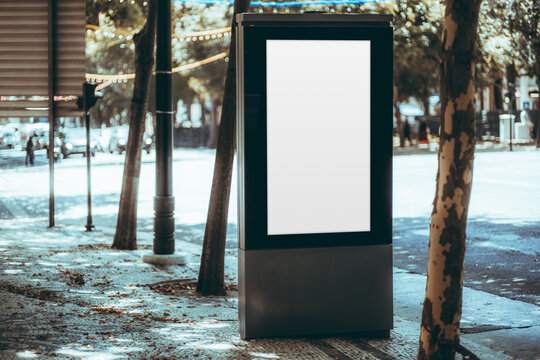 Vertical advertising template in a freestanding urban lightbox, surrounded by leafy trees, patterned pavement, string lights and a softly blurred city street
