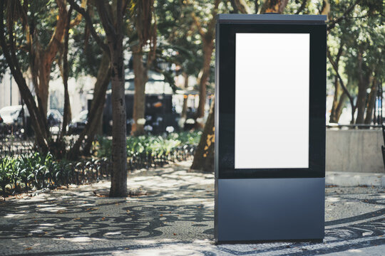 Blank vertical advertising template on a freestanding lightbox beside a leafy promenade, with patterned mosaic pavement and generous copy space on the left