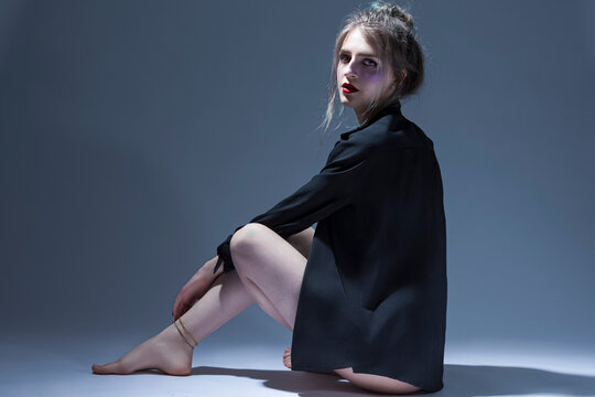 Violence and Harassment Ideas. Young Caucasian Girl in Violet Nightie and Black Shirt Sitting and Posing on Gray Background With Bruses Under Eyes on Face.