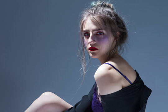 Violence and Harassment Ideas. Young Caucasian Girl in Violet Nightie and Black Shirt Sitting and Posing on Gray Background With Bruses Under Eyes on Face.