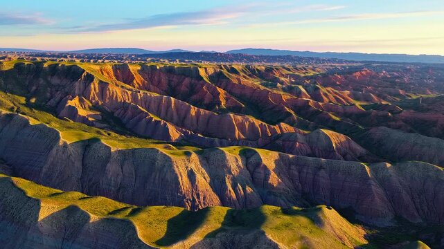 Aerial view of dramatic multi-colored mountain ridges and deep canyons illuminated by warm sunlight under a soft twilight sky.