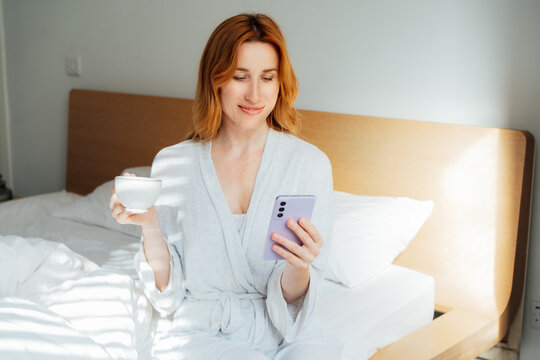 Smiling Middle aged woman in pajama scrolling phone and drinking morning coffee in her bed. Modern morning routine and habits. Work at home. Morning news, social media, blog, surfing on internet