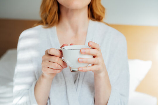 Close up female hands holding cup of coffee relaxing in bed in morning sunlight. Hot drink for wake up and energy boost. Well being morning routine and daydreaming mood. Good morning habits.