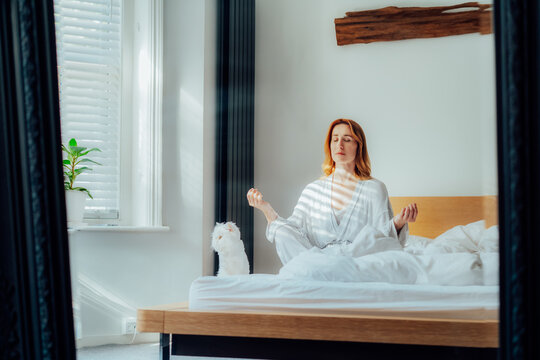 Mirror reflection of woman in pajama and white cat pet meditating on bed, practicing mindfulness and relaxation for a peaceful morning self-care and wellness routine at home. Mental health practice.