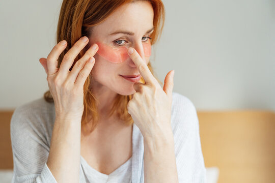 Self Cosmetology, skin care, face treatment, spa, natural beauty concept. Smiling middle aged woman in pajamas with mirror applying pink moisturizing patches on eyes. Home Beauty routine.