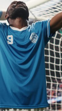 Vertical screen: African American striker kissing ground for football celebration after scoring goal and winning match, goal scorer celebrating victory and kneeling on pitch.