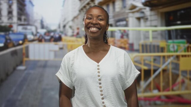 Young african american woman smiling showing teeth, standing relaxed in a city street beside construction barriers and yellow railings with a casual white buttoned shirt; joy confidence.