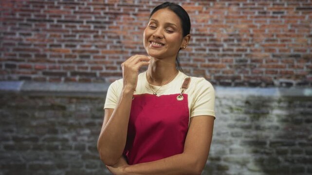 Young hispanic brunette woman in red apron touches cheek with hand while smiling in front of a brick building wall in casual service pose; warmth hospitality.