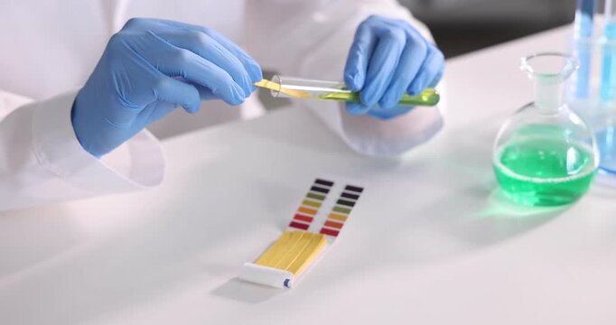 Scientist holding PH test strip and glass tube with liquid at white table in laboratory, closeup